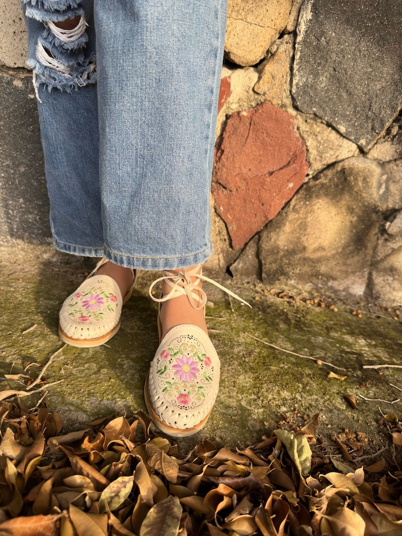 May include: Cream-colored leather sandals with floral embroidery in pink, green, and purple. The sandals have a woven design and ankle ties. The person is wearing light blue ripped jeans. The background is a stone wall and fallen leaves.