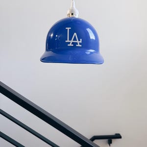 May include: A blue pendant light shaped like a baseball cap, featuring the white letters "LA". The light hangs from a white cord against a white wall. The light fixture is a unique home decor item.