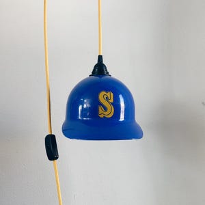 May include: A blue baseball cap shaped pendant light with a gold 'S' on the front. The light is hanging from a cord with a black switch.