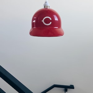 May include: A red baseball cap-shaped pendant light with a white "C" on the front, hanging from a white cord against a white wall.