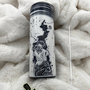 May include: A white tumbler with a black and white design of a woman standing in front of a starry sky. The tumbler has a black lid and a black and white glitter design around the top. The tumbler is sitting on a white fuzzy surface. The text "Lunatic's Creation" is visible on a sticker.