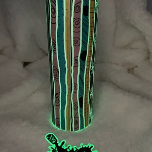 Op de afbeelding: Een gloeiende beker met een gestreept patroon in tinten turquoise, roze, zwart en goud. Het patroon heeft een herhalend ontwerp van swirls en lijnen. De beker heeft een zwart deksel en een witte sticker met de tekst "Lunatic's Creation" in zwart.