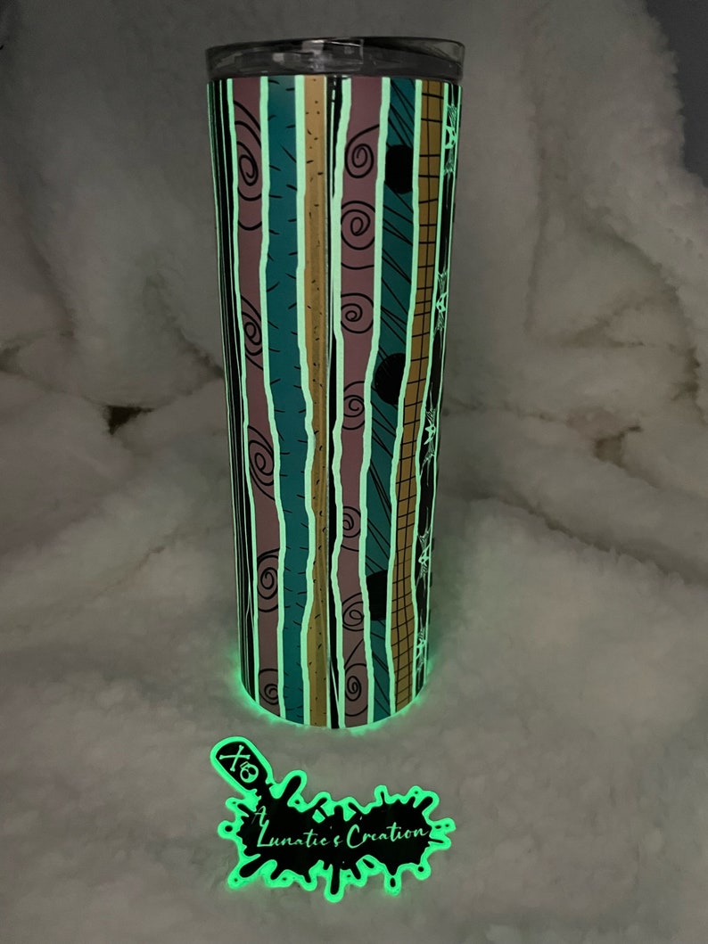 Op de afbeelding: Een gloeiende beker met een gestreept patroon in tinten turquoise, roze, zwart en goud. Het patroon heeft een herhalend ontwerp van swirls en lijnen. De beker heeft een zwart deksel en een witte sticker met de tekst "Lunatic's Creation" in zwart.