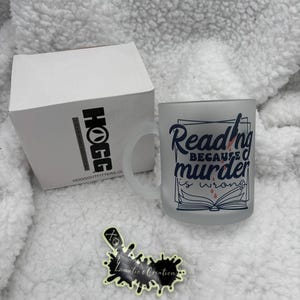 Może przedstawiać: Biały kubek ceramiczny z niebiesko-białym wzorem, na którym widnieje napis "Reading because murder is wrong". Na książce widoczne są czerwone krople krwi.