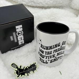 May include: White ceramic mug with a black rim and handle. The mug has black text that reads "Running on low tire pressure and five miles till empty".