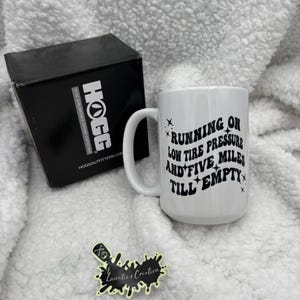 May include: White ceramic coffee mug with a black design that reads "Running on low tire pressure and five miles till empty". The mug is sitting on a white fluffy surface.