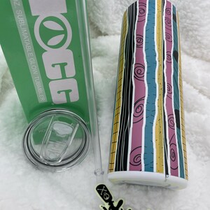 Op de afbeelding: Een 20 oz. sublimatierende glow-beker met een gestreept ontwerp in zwart, wit, roze, blauw en geel. De beker heeft een doorzichtig deksel en een rietje. De beker staat op een wit, pluche oppervlak. De tekst "HOGG" is afgedrukt op de groene doos naast de beker.