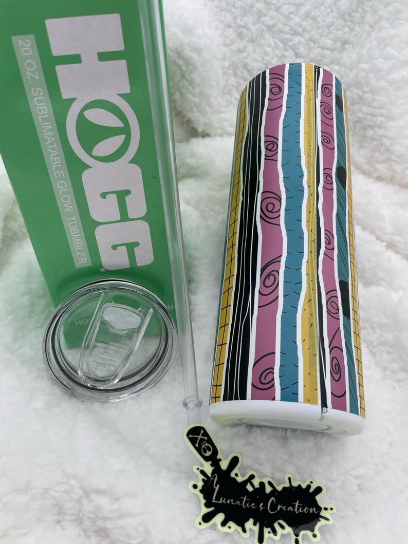Op de afbeelding: Een 20 oz. sublimatierende glow-beker met een gestreept ontwerp in zwart, wit, roze, blauw en geel. De beker heeft een doorzichtig deksel en een rietje. De beker staat op een wit, pluche oppervlak. De tekst "HOGG" is afgedrukt op de groene doos naast de beker.