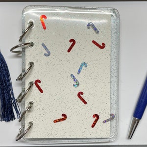 Könnte beinhalten: Ein weißes Glitzer-Notizbuch mit einem durchsichtigen Cover und silbernen Ringen. Das Cover ist mit roten und silbernen Zuckerstangen-Formen verziert. Das Notizbuch wird mit einem passenden Lesezeichen und einem blauen Stift mit rotem Glitzer im Inneren geliefert.