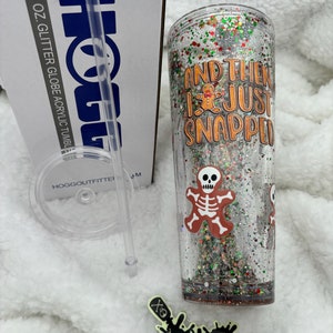 May include: A clear 24 ounce glitter globe acrylic tumbler with a red, green, and white glitter base. The tumbler has a white lid and a clear straw. The tumbler has a gingerbread man and skeleton graphic with the text "And then I just snapped".