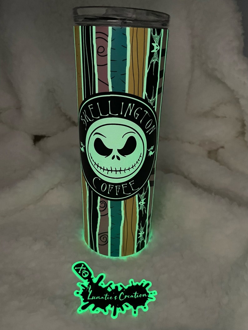 Op de afbeelding: Een glow-in-the-dark tumbler met een zwart-wit ontwerp met Jack Skellington uit The Nightmare Before Christmas. De tumbler heeft een zwarte cirkel met de tekst "Skellington Coffee" en een afbeelding van Jack Skellingtons gezicht. De tumbler is omgeven door een witte achtergrond.