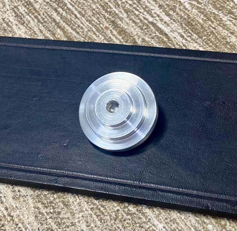 Peut inclure: Petit objet m&eacute;tallique argent&eacute; de forme circulaire, perc&eacute; d'un trou central. L'objet pr&eacute;sente un aspect &eacute;tag&eacute;, avec des anneaux concentriques cr&eacute;ant un effet de paliers. Il repose sur une surface sombre et textur&eacute;e.