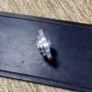 Peut inclure: Un objet m&eacute;tallique argent&eacute; brillant en forme d'&eacute;toile, avec plusieurs pointes et une vis centrale. L'objet est pos&eacute; sur un fond sombre et textur&eacute;, peut-&ecirc;tre du cuir ou un mat&eacute;riau similaire.