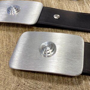 Peut inclure: Deux boucles de ceinture en m&eacute;tal argent&eacute; avec une finition bross&eacute;e et un motif circulaire superpos&eacute; au centre. Les boucles sont fix&eacute;es &agrave; des ceintures en cuir noir. Le fond est une surface textur&eacute;e brun clair.