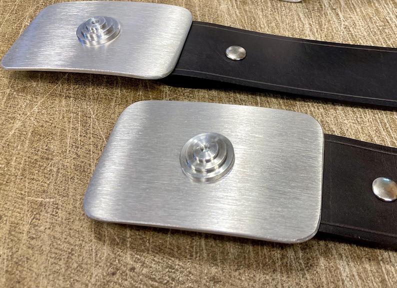 Peut inclure: Deux boucles de ceinture en m&eacute;tal argent&eacute; avec une finition bross&eacute;e et un motif circulaire superpos&eacute; au centre. Les boucles sont fix&eacute;es &agrave; des ceintures en cuir noir. Le fond est une surface textur&eacute;e brun clair.