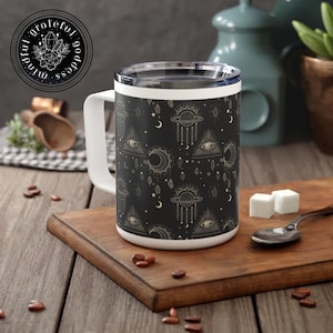 May include: A white ceramic mug with a black background and gold celestial pattern. The pattern includes moons, stars, planets, eyes, and crystals. The mug has a handle and a white rim.