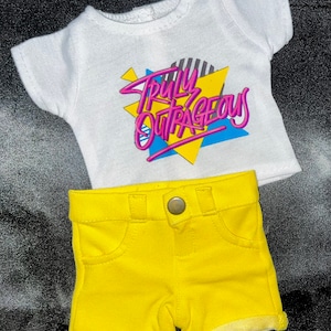 May include: A white t-shirt with short sleeves and the text "Truly Outrageous" in pink and blue letters on a yellow and blue geometric design. The shirt is paired with yellow denim shorts.