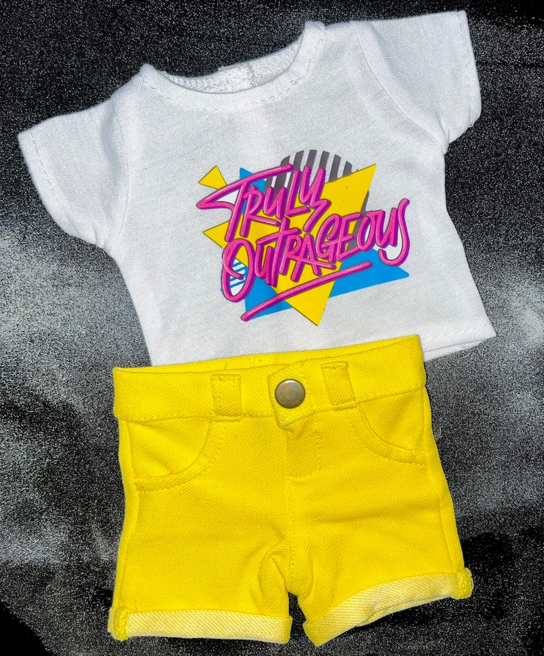 May include: A white t-shirt with short sleeves and the text "Truly Outrageous" in pink and blue letters on a yellow and blue geometric design. The shirt is paired with yellow denim shorts.