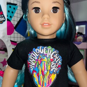 May include: A doll with dark hair featuring bright blue highlights, wearing a black short-sleeved t-shirt. The shirt displays a colorful graphic print of a person with the text "UNAPOLOGETICALLY DOPE" arched over the top in white lettering.