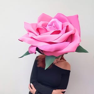 Puede incluir: Una mujer con un gran tocado de rosa rosa y un vestido negro con hombros descubiertos. El vestido tiene mangas largas y detalles de encaje. Lleva sandalias negras.