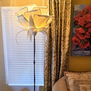 May include: A unique floor lamp with a large, white, flower-shaped shade. The lamp has a black metal pole and base. The shade is lit from within, casting a warm glow. The lamp is positioned near a window with blinds and a patterned curtain.