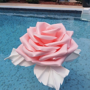 May include: A large, pink rose pool float with white leaves floats on the surface of a blue swimming pool. The rose has detailed petals and appears to be made of a soft material. The water has a slight ripple.