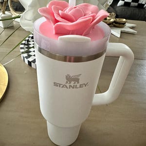 Peut inclure: Un mug de voyage blanc Stanley avec une poign&eacute;e, surmont&eacute; d'un couvercle en forme de rose rose et d'une paille blanche. Le mug a un bord argent&eacute; et le logo Stanley. Le mug est sur une surface marron clair.