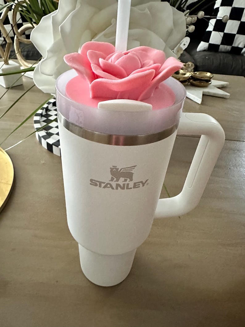 Peut inclure: Un mug de voyage blanc Stanley avec une poign&eacute;e, surmont&eacute; d'un couvercle en forme de rose rose et d'une paille blanche. Le mug a un bord argent&eacute; et le logo Stanley. Le mug est sur une surface marron clair.