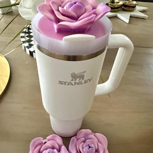 Peut inclure: Mug de voyage blanc de la marque Stanley avec une poign&eacute;e, un couvercle rose et une paille blanche. Le mug est surmont&eacute; d'une grande fleur d&eacute;corative violette. Deux d&eacute;corations de fleurs violettes suppl&eacute;mentaires sont &agrave; la base du mug.