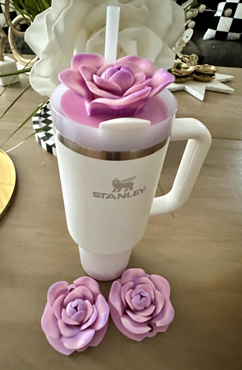 Peut inclure: Mug de voyage blanc de la marque Stanley avec une poign&eacute;e, un couvercle rose et une paille blanche. Le mug est surmont&eacute; d'une grande fleur d&eacute;corative violette. Deux d&eacute;corations de fleurs violettes suppl&eacute;mentaires sont &agrave; la base du mug.