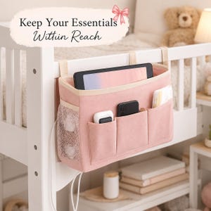 May include: Pink bedside organizer with multiple pockets, hanging from a white bed frame. The organizer holds a tablet, phone, and other items. The text "Keep Your Essentials Within Reach" is displayed above the organizer.