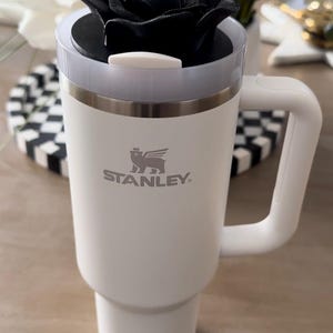 Peut inclure: Mug de voyage Stanley blanc avec un couvercle en forme de rose noire et une paille blanche. Le mug a une poign&eacute;e et le logo Stanley. Le mug est sur une surface en bois.