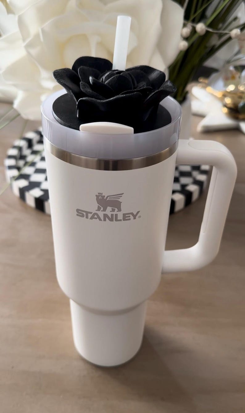 Peut inclure: Mug de voyage Stanley blanc avec un couvercle en forme de rose noire et une paille blanche. Le mug a une poign&eacute;e et le logo Stanley. Le mug est sur une surface en bois.