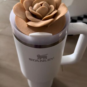 Peut inclure: Mug de voyage Stanley blanc avec un couvercle en forme de fleur marron clair et une paille blanche. La tasse a une poign&eacute;e et le logo Stanley est visible. Le couvercle en forme de fleur ajoute une touche d&eacute;corative.