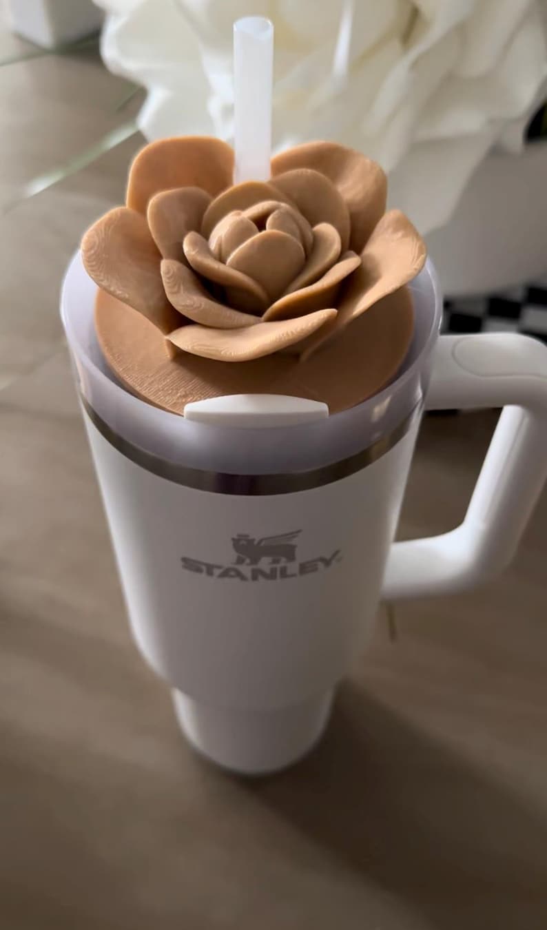 Peut inclure: Mug de voyage Stanley blanc avec un couvercle en forme de fleur marron clair et une paille blanche. La tasse a une poign&eacute;e et le logo Stanley est visible. Le couvercle en forme de fleur ajoute une touche d&eacute;corative.