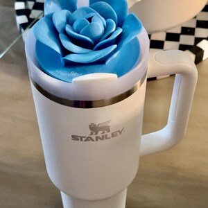 Peut inclure: Gobelet Stanley blanc avec un couvercle bleu en forme de fleur et une poign&eacute;e blanche. Le gobelet porte le logo Stanley sur le devant. Une paille blanche est ins&eacute;r&eacute;e dans le couvercle. Le gobelet est sur une surface en bois.