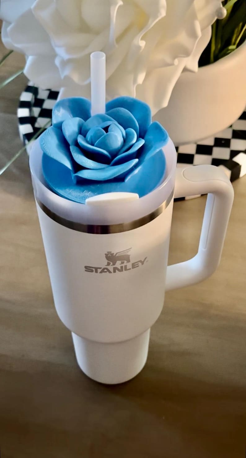 Peut inclure: Gobelet Stanley blanc avec un couvercle bleu en forme de fleur et une poign&eacute;e blanche. Le gobelet porte le logo Stanley sur le devant. Une paille blanche est ins&eacute;r&eacute;e dans le couvercle. Le gobelet est sur une surface en bois.