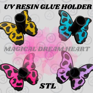 May include: Four butterfly-shaped UV resin glue holders in yellow, blue, pink, and purple. Each holder has a black tube for glue and black detailing on the wings. The text "UV RESIN GLUE HOLDER", "MAGICAL DREAM HEART", and "STL" are visible.