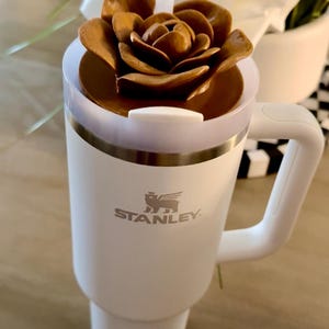 Peut inclure: Un mug de voyage blanc Stanley avec une poign&eacute;e, surmont&eacute; d'un couvercle marron en forme de fleur. Une paille blanche d&eacute;passe du couvercle. Le logo Stanley est visible &agrave; l'avant du mug.