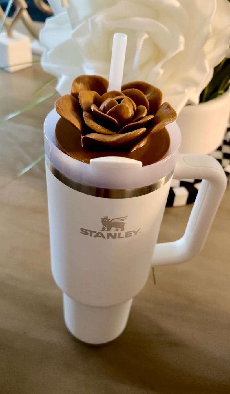 Peut inclure: Un mug de voyage blanc Stanley avec une poign&eacute;e, surmont&eacute; d'un couvercle marron en forme de fleur. Une paille blanche d&eacute;passe du couvercle. Le logo Stanley est visible &agrave; l'avant du mug.