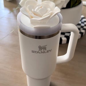 Peut inclure: Mug de voyage blanc Stanley avec un couvercle en forme de fleur blanche et une paille transparente. Le mug a une poign&eacute;e et le logo Stanley. Le mug est sur une surface claire.