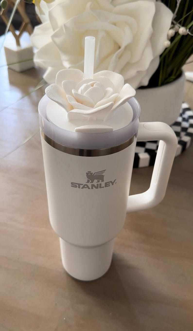 Peut inclure: Mug de voyage blanc Stanley avec un couvercle en forme de fleur blanche et une paille transparente. Le mug a une poign&eacute;e et le logo Stanley. Le mug est sur une surface claire.