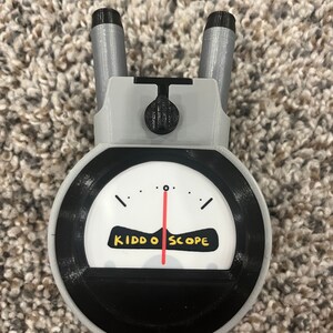 Op de afbeelding: Een grijze en zwarte Kiddo Scope met een rode naald op een witte wijzerplaat. De wijzerplaat heeft zwarte markeringen en de tekst "Kiddo Scope" in geel.