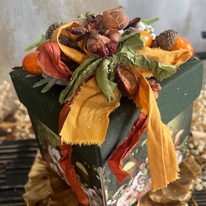 Può includere: Una scatola decorativa verde scuro con una composizione floreale. La composizione include bacche arancioni, fiori secchi e nastri verdi e arancioni. La scatola ha un disegno stampato sui lati.