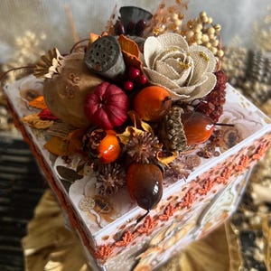 Può includere: Una scatola decorativa a tema autunnale, adornata con una varietà di elementi autunnali. La composizione include un fiore di juta, zucche in miniatura, ghiande e bacche. La scatola è decorata con un motivo floreale e bordi in pizzo arancione.
