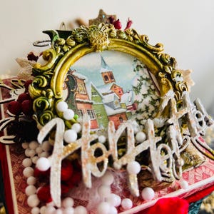May include: A decorative Christmas-themed frame with a winter village scene inside. The frame is gold with ornate detailing and the word "Ho Ho Ho" in white and gold. Red berries and white spheres add festive accents.