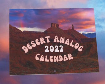 May include: A rectangular wall calendar featuring a landscape photograph of a desert rock formation under a sunset sky with pink and purple clouds. The text "DESERT ANALOG 2022 CALENDAR" is printed in a light pink, rounded, retro-style font.