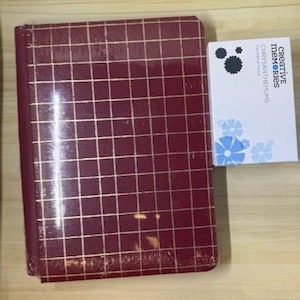 May include: A burgundy photo album with a gold grid pattern. Next to it is a white box with the text "Creative Memories" and "Chrysanthemums" with blue and black floral designs. The album is rectangular and appears to be new.