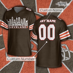 以下が含まれることがあります： 袖に白とオレンジのストライプが入った茶色のフットボールジャージ。前面には、選手名と「クリーブランド」のスカイライングラフィックがあります。背面には番号「00」とカスタム名用のスペースがあります。
