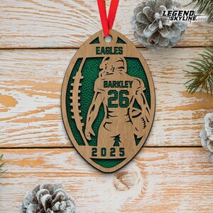 May include: Wooden football-shaped ornament with a green interior and a player silhouette. The ornament features the word "EAGLES" at the top, "BARKLEY 26" in the center, and "2025" at the bottom. A red ribbon is attached for hanging.